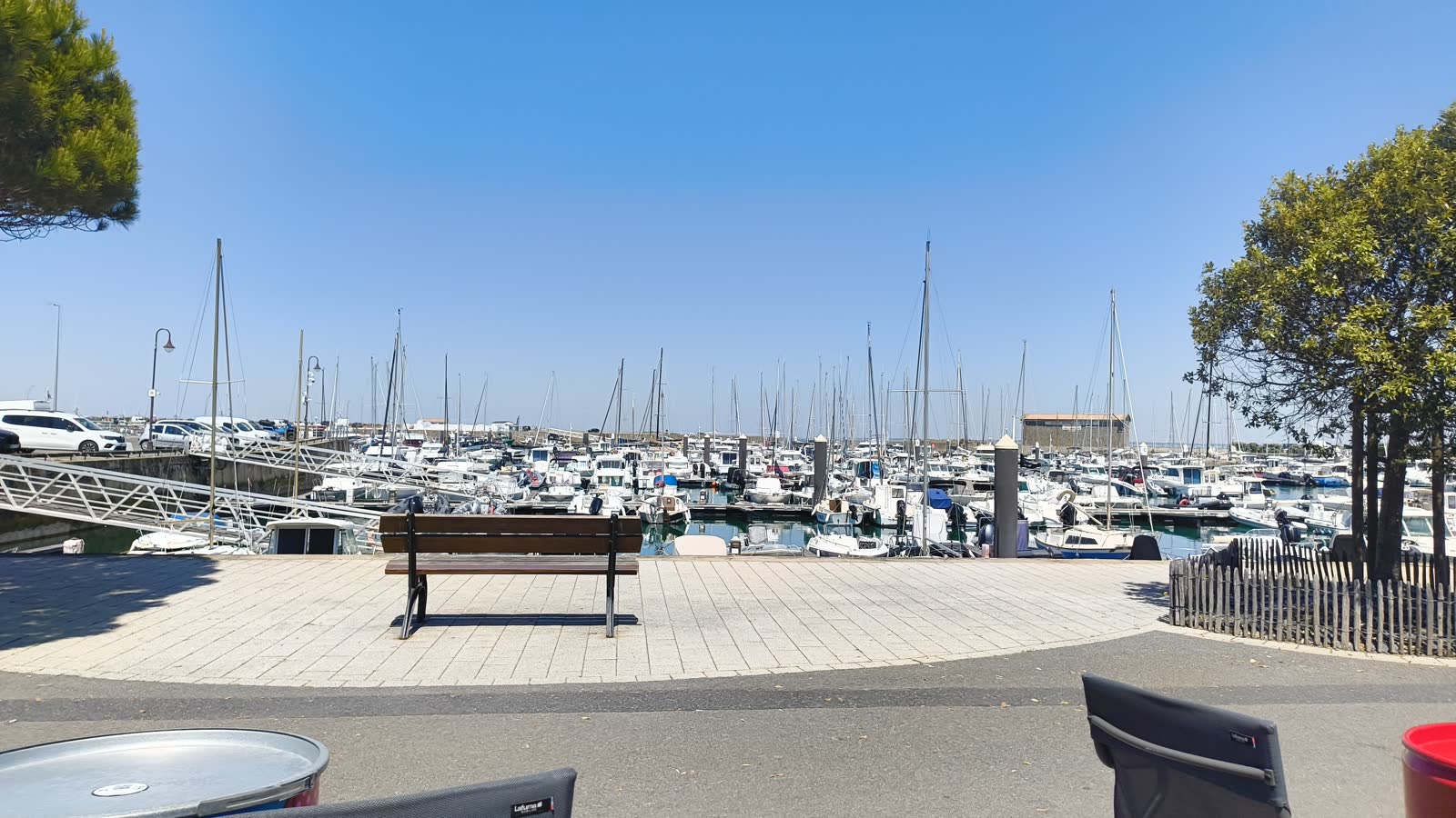 Port de Noirmoutier avec des bateaux amarrés, des arbres, un banc et la moto visible au premier plan.